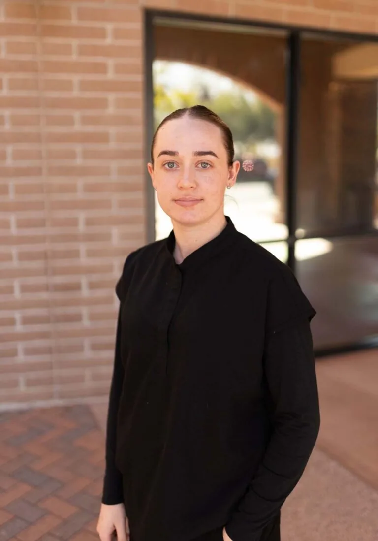 A woman wearing a black shirt standing in front of a brick of wall with one of her hand in the pocket.