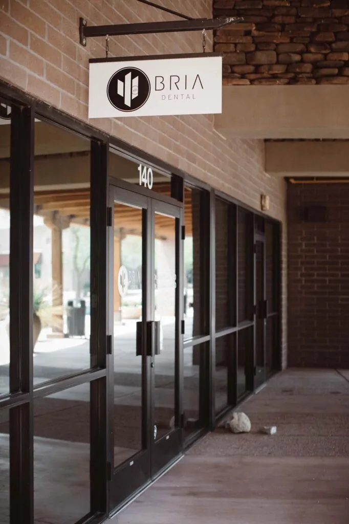 Exterior view of Bria Dental, located in a brick-facade commercial building with large glass windows and doors. The office features a hanging sign with the 'Bria Dental' logo above the entrance, which is marked with the number '140'.