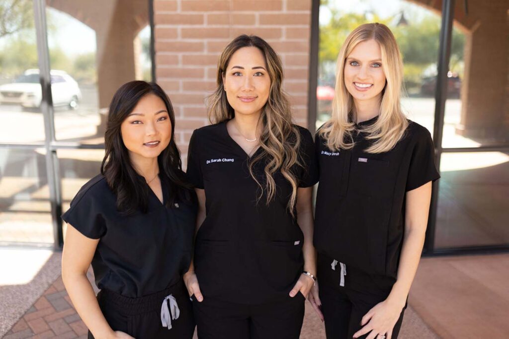 Three woman standing together in front of a brick wall, wearing a black scrubs.
