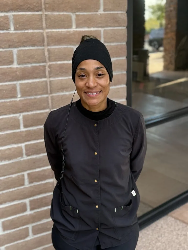 Person standing in front of a brick wall, wearing a black beanie, black jacket with buttons, and black pants. The wire or cord is visible on the jacket, possibly connecting to a device.